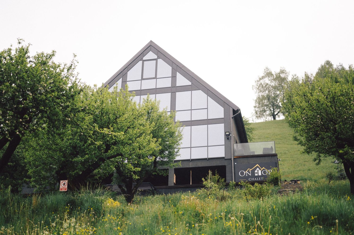 A modern A-frame house with white and gray exterior stands on a grassy hillside surrounded by green trees and bushes.