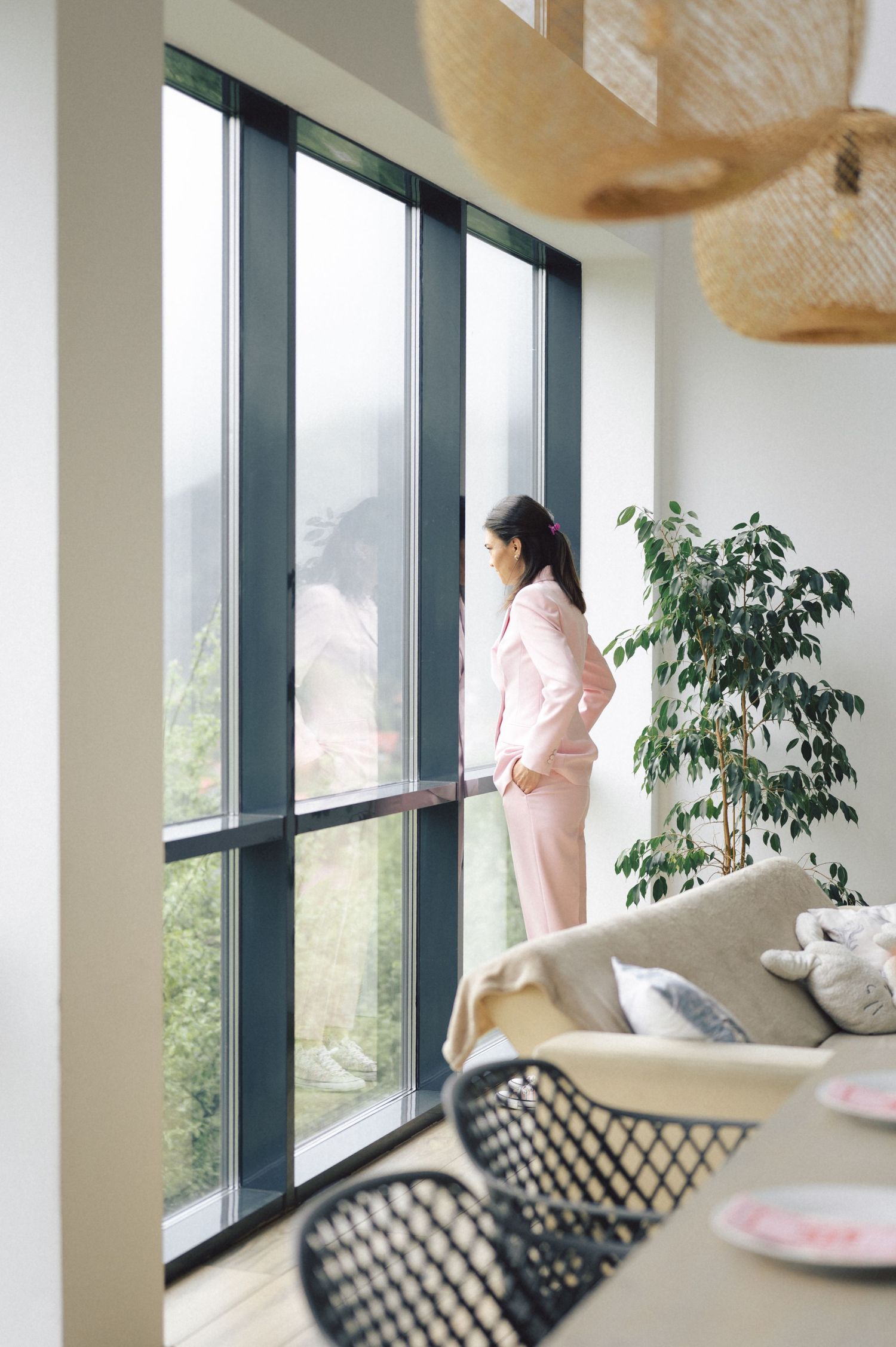 Someone in pink loungewear looks out floor-to-ceiling windows in a modern room with contemporary decor and plants.