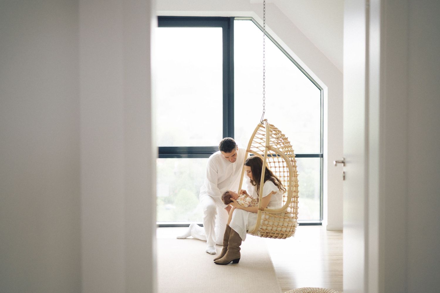 Someone relaxes in a hanging rattan egg chair by a bright window in a minimalist white interior space.