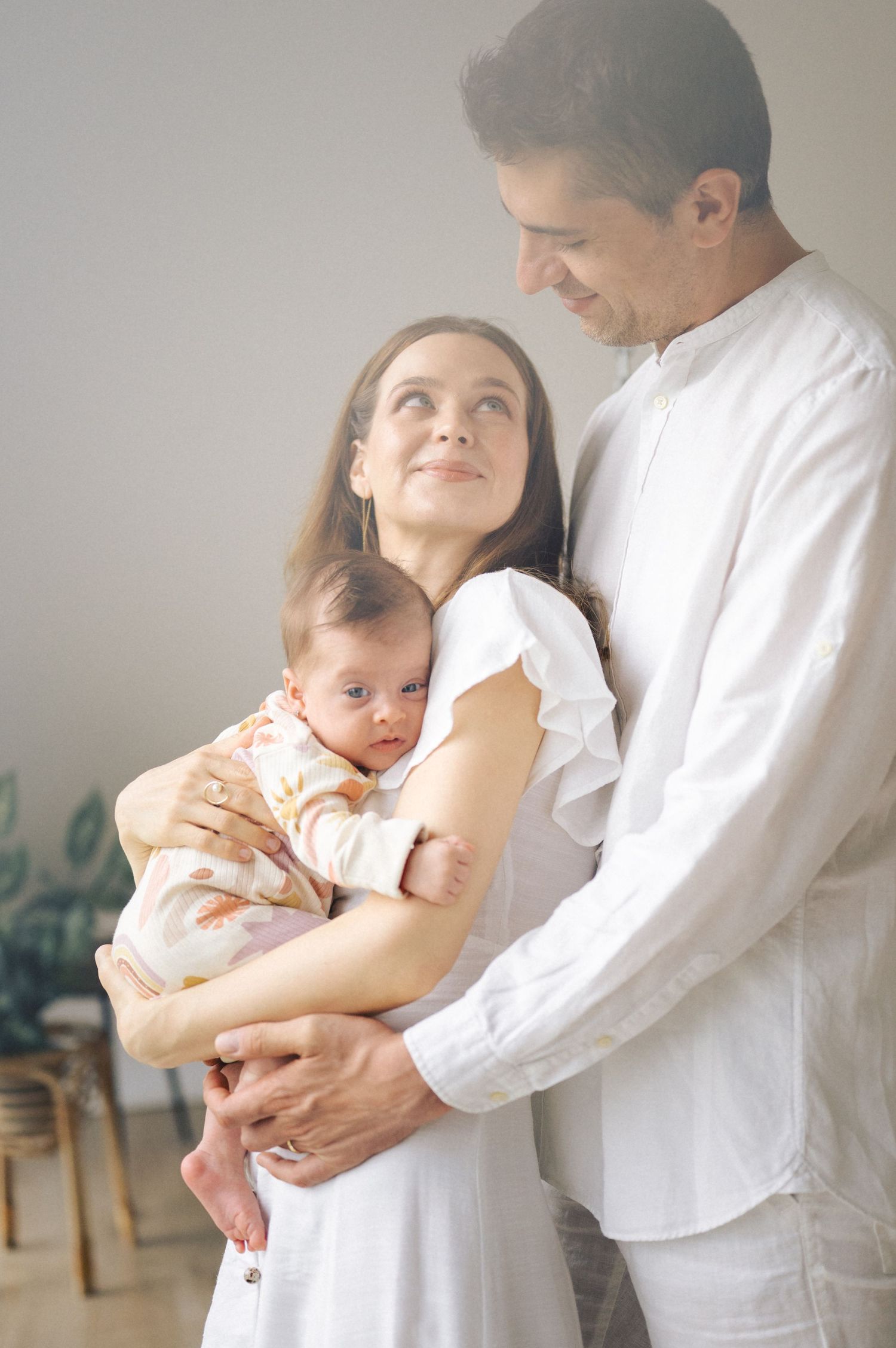 A couple in white clothing share a tender moment while holding their infant child in a softly lit room.