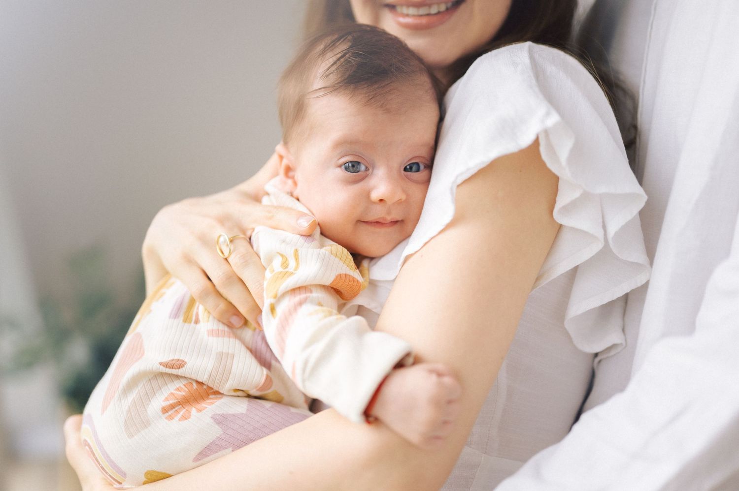A baby in a cream-colored outfit is being held lovingly in someone's arms against a light background.