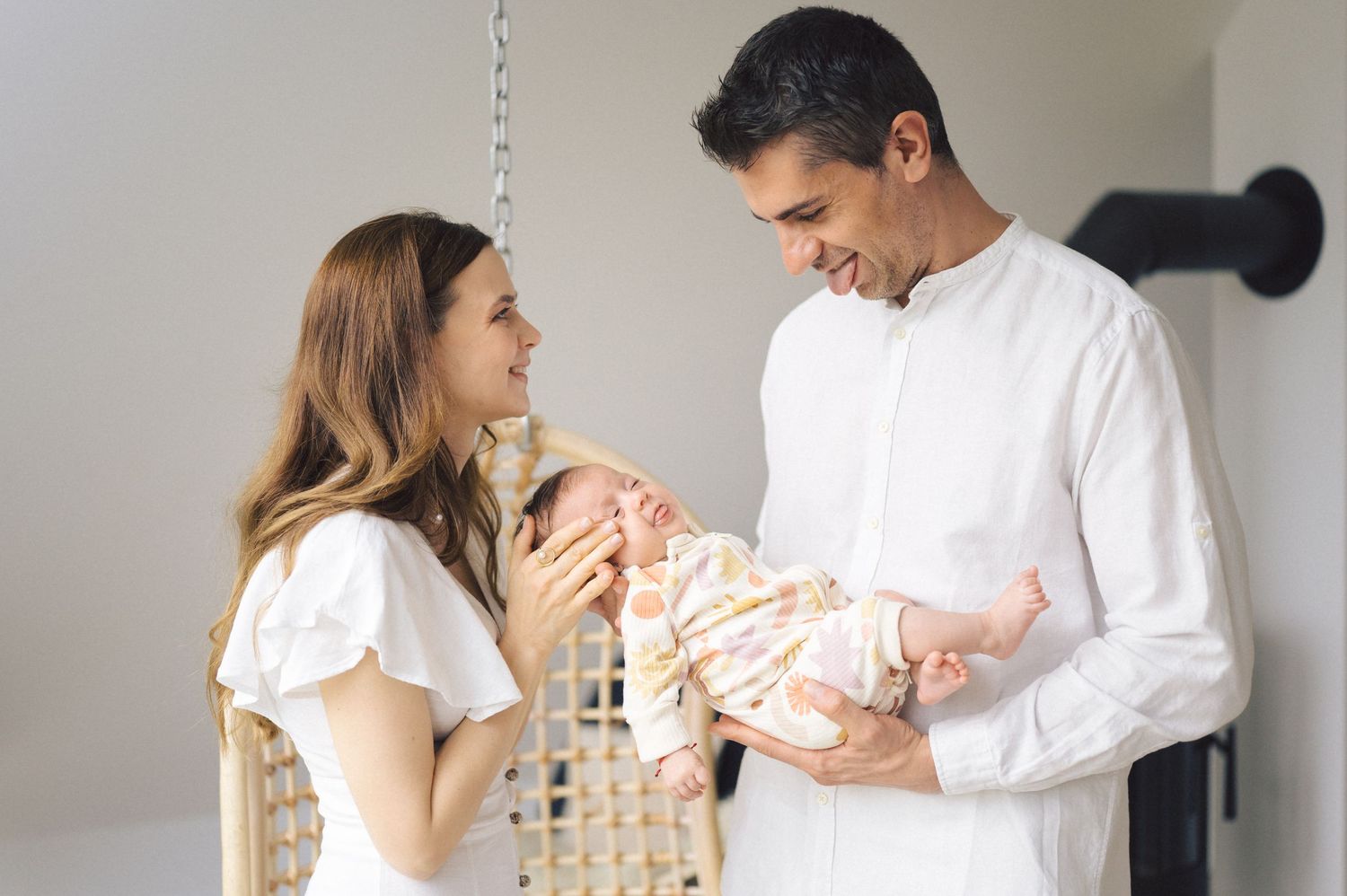A couple stands together in a white room, lovingly gazing at a swaddled newborn baby in their arms.