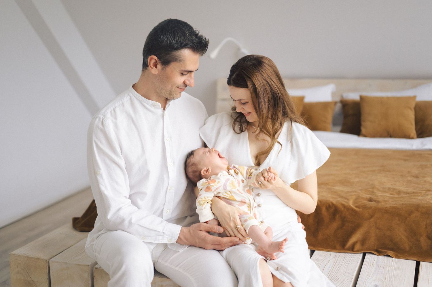 Parents wearing white outfits sit together on a bedroom floor while lovingly holding their newborn baby.