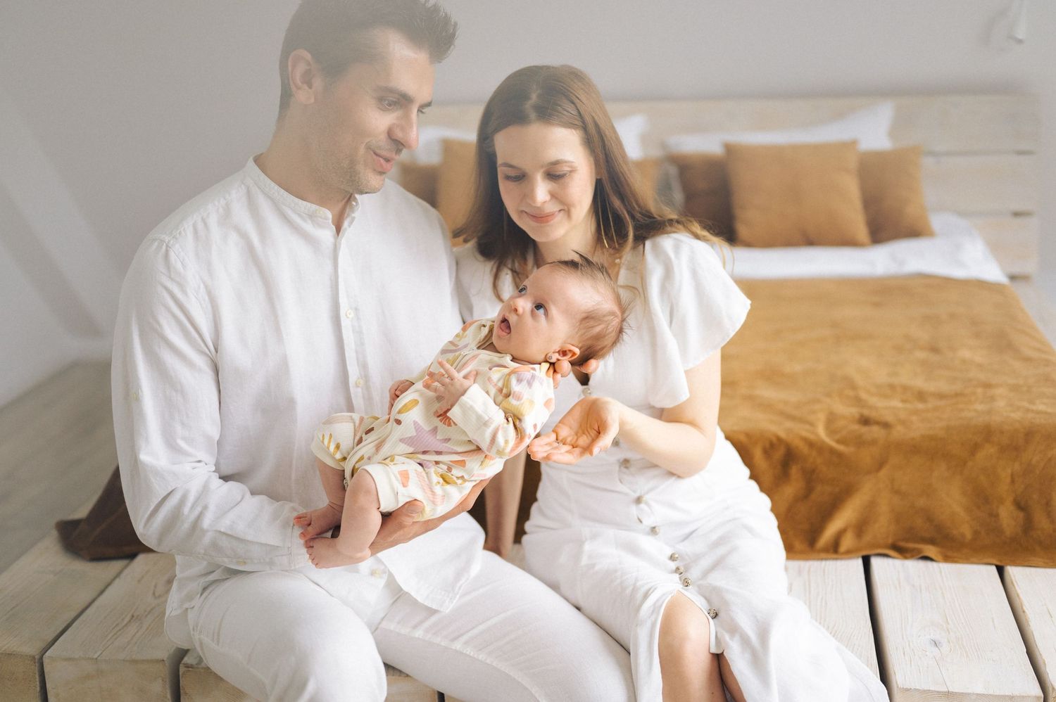 Happy family in white clothing sitting together on wooden floor in bright bedroom with infant baby.