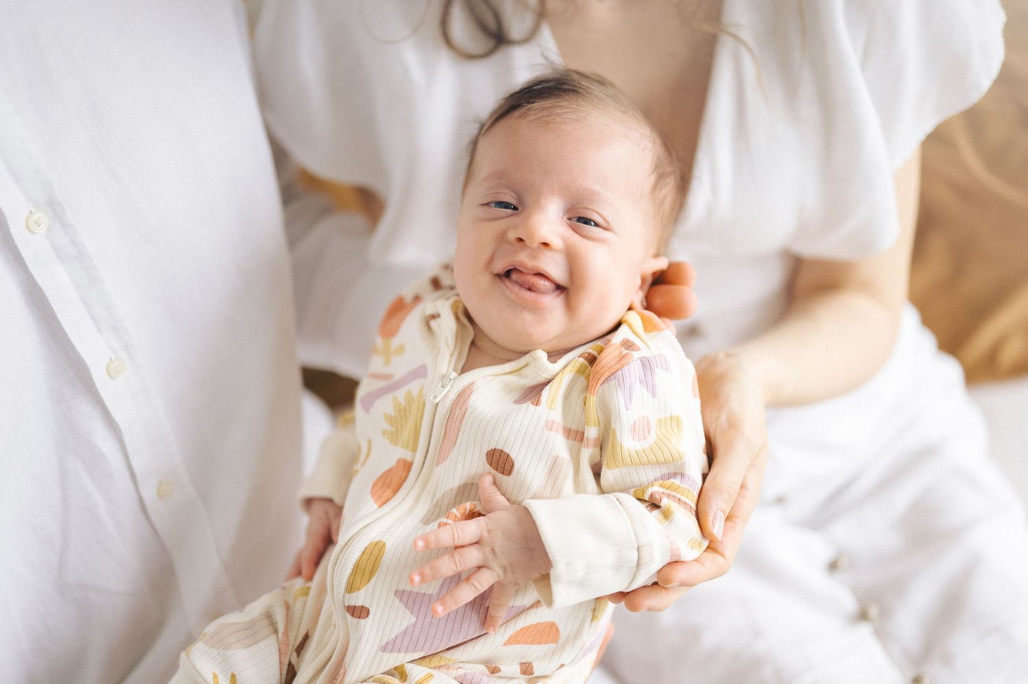 Baby wearing a peach and white patterned dress smiles while being held against bright white fabric.