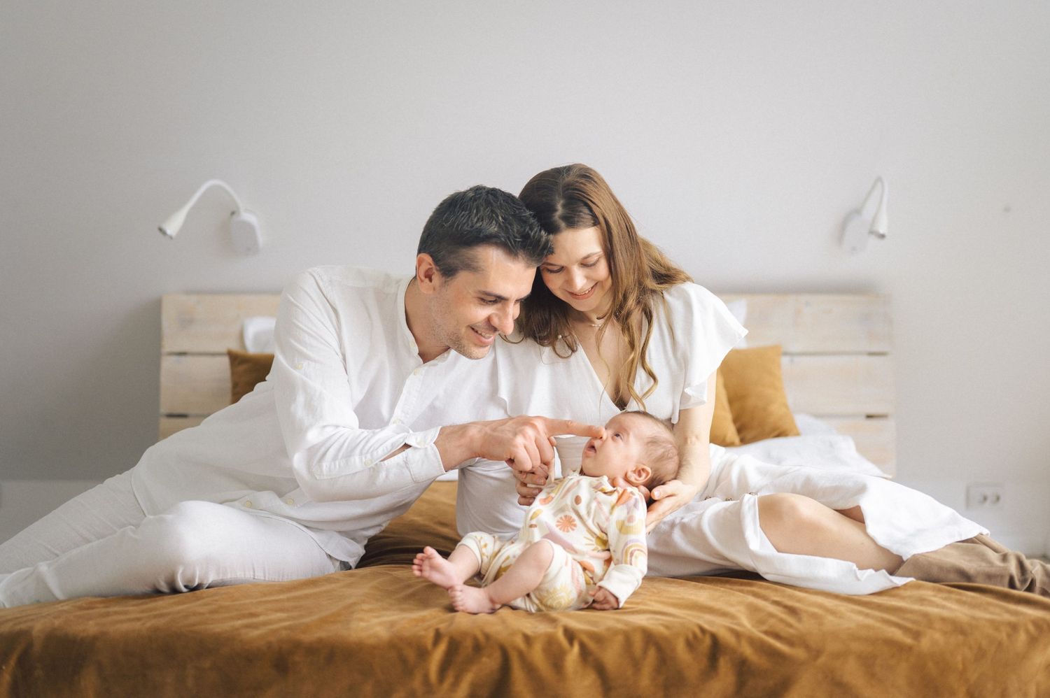 Parents in white pajamas lovingly playing with their newborn baby on a brown bed with white bedding and teddy bear.
