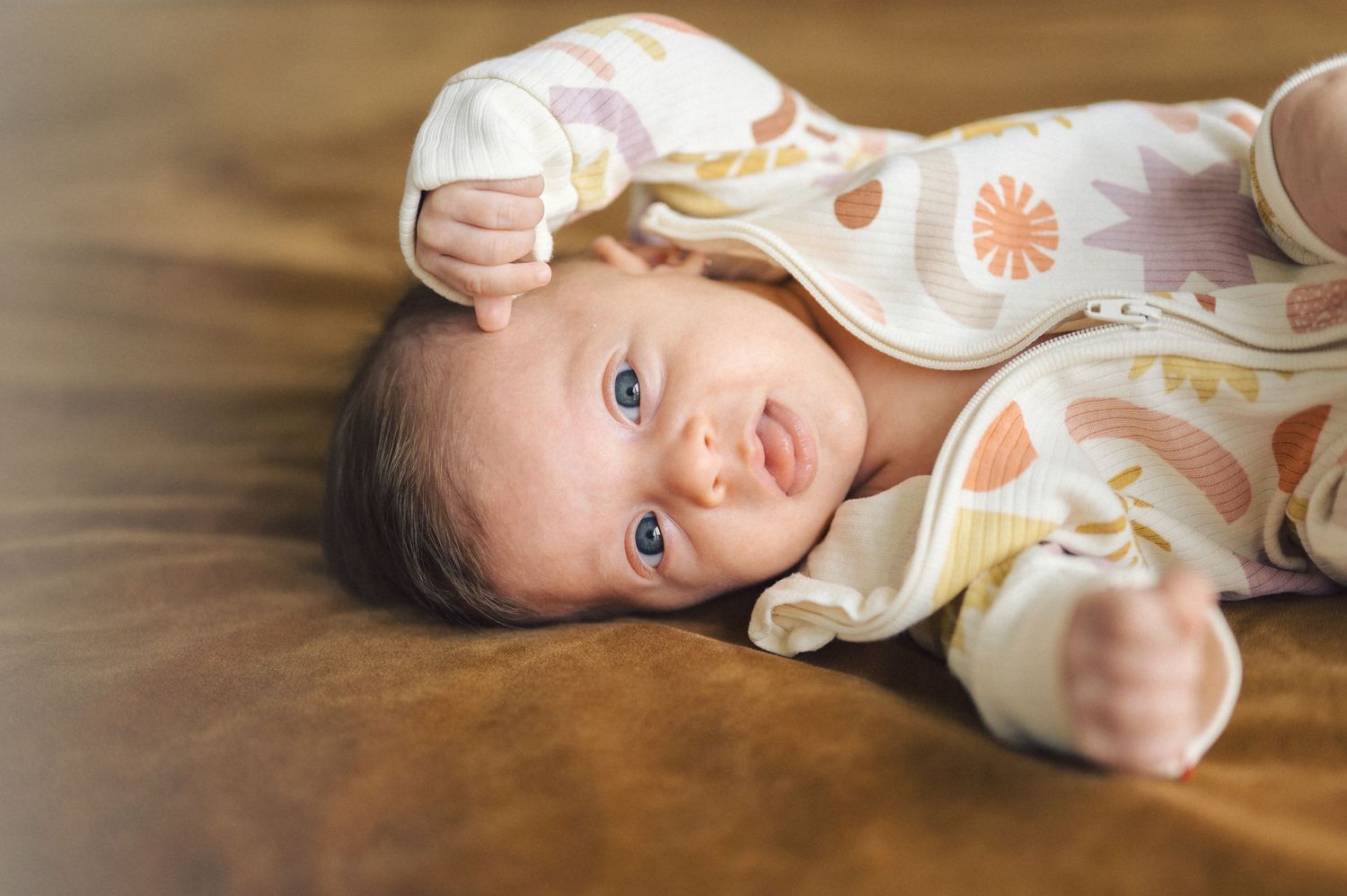 A baby wearing a floral patterned onesie lies on a wooden floor while smiling.
