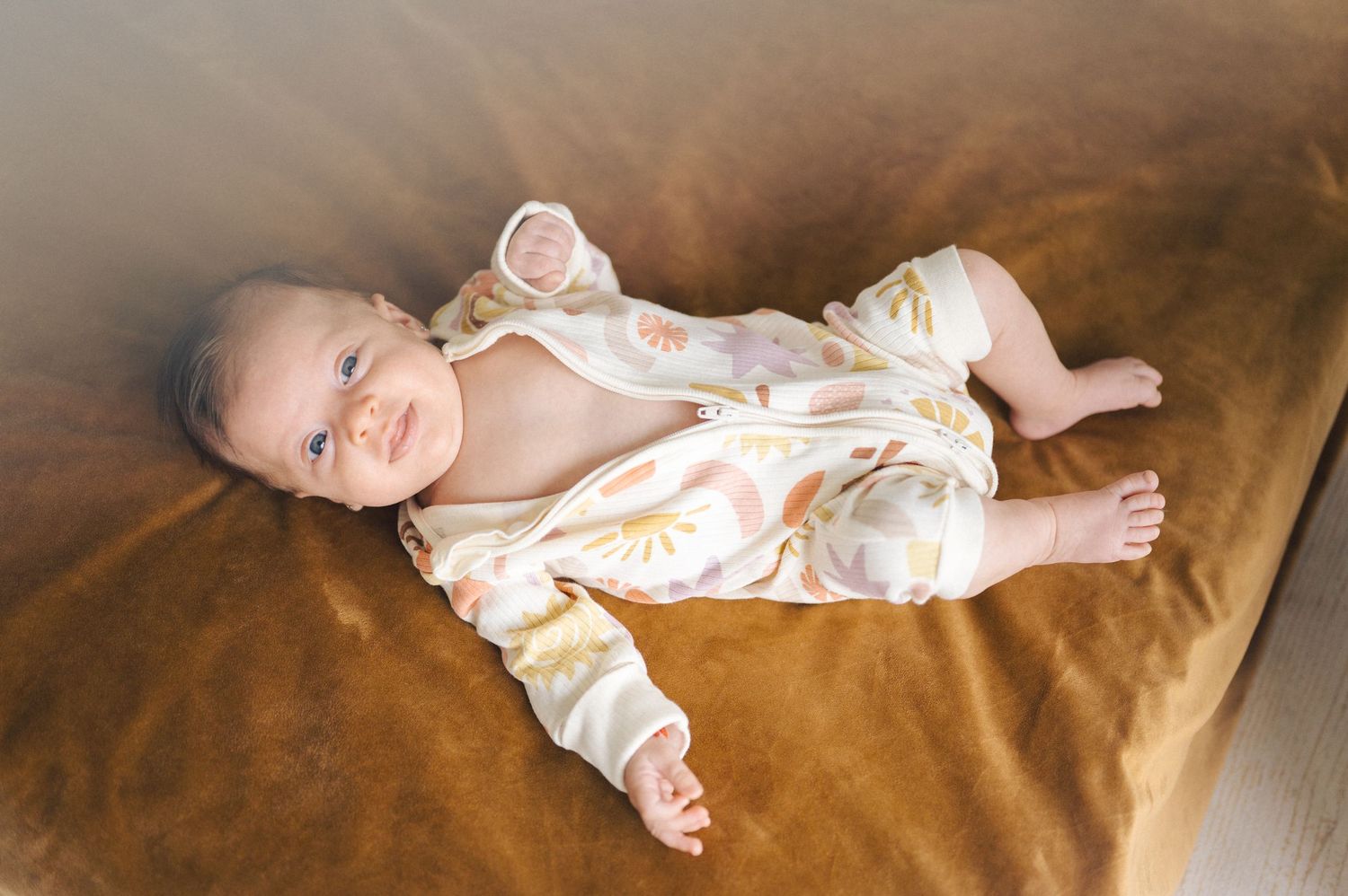A baby wearing floral sleepwear smiles while lying on a brown velvet couch.