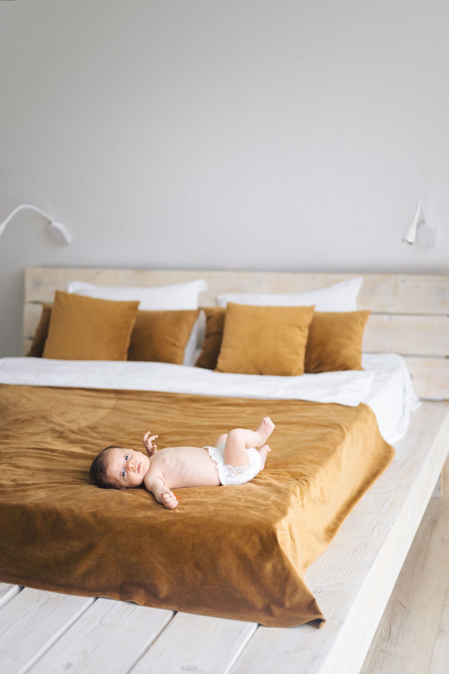Baby in pink onesie lying on rustic bed with white sheets and mustard yellow throw pillows and blanket.
