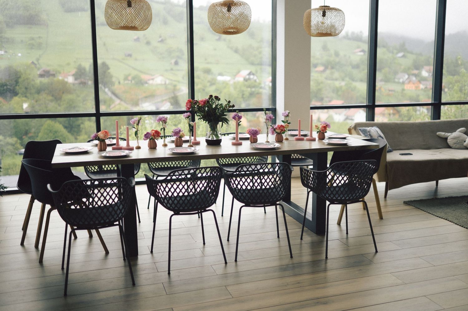 Elegant dining table set with black chairs and pendant lights in modern room with panoramic windows overlooking scenery.