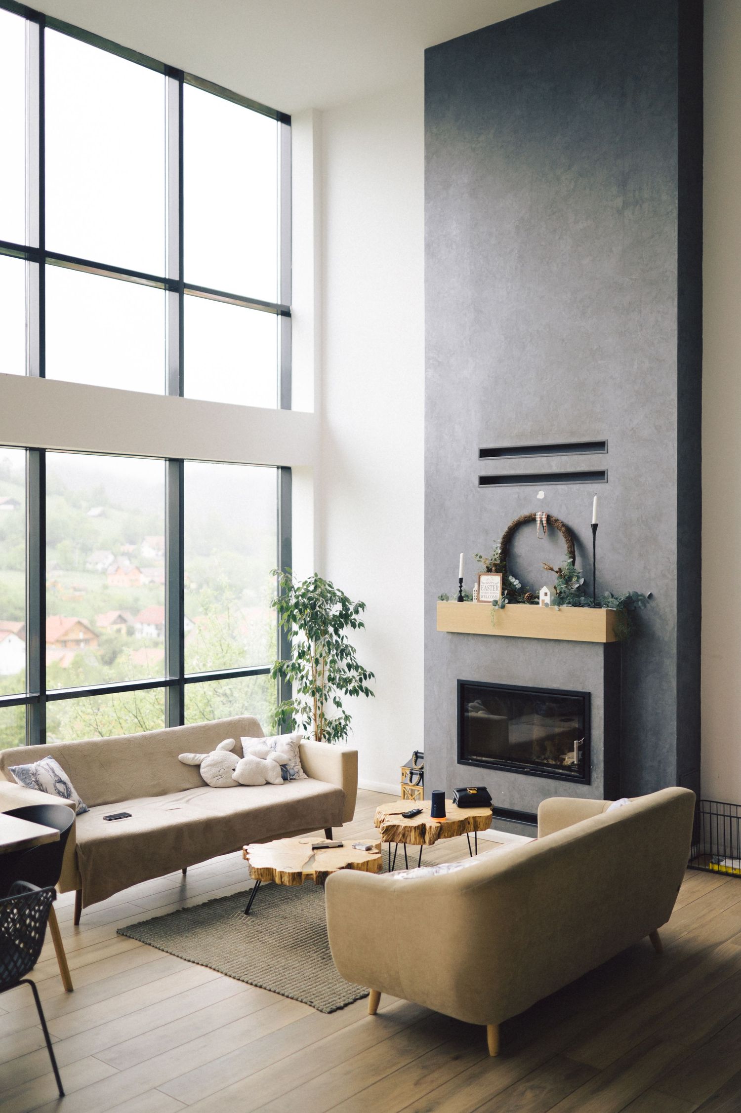 Modern living room with floor-to-ceiling windows, tall grey fireplace, tan sofas, and hardwood floors in a minimalist style.