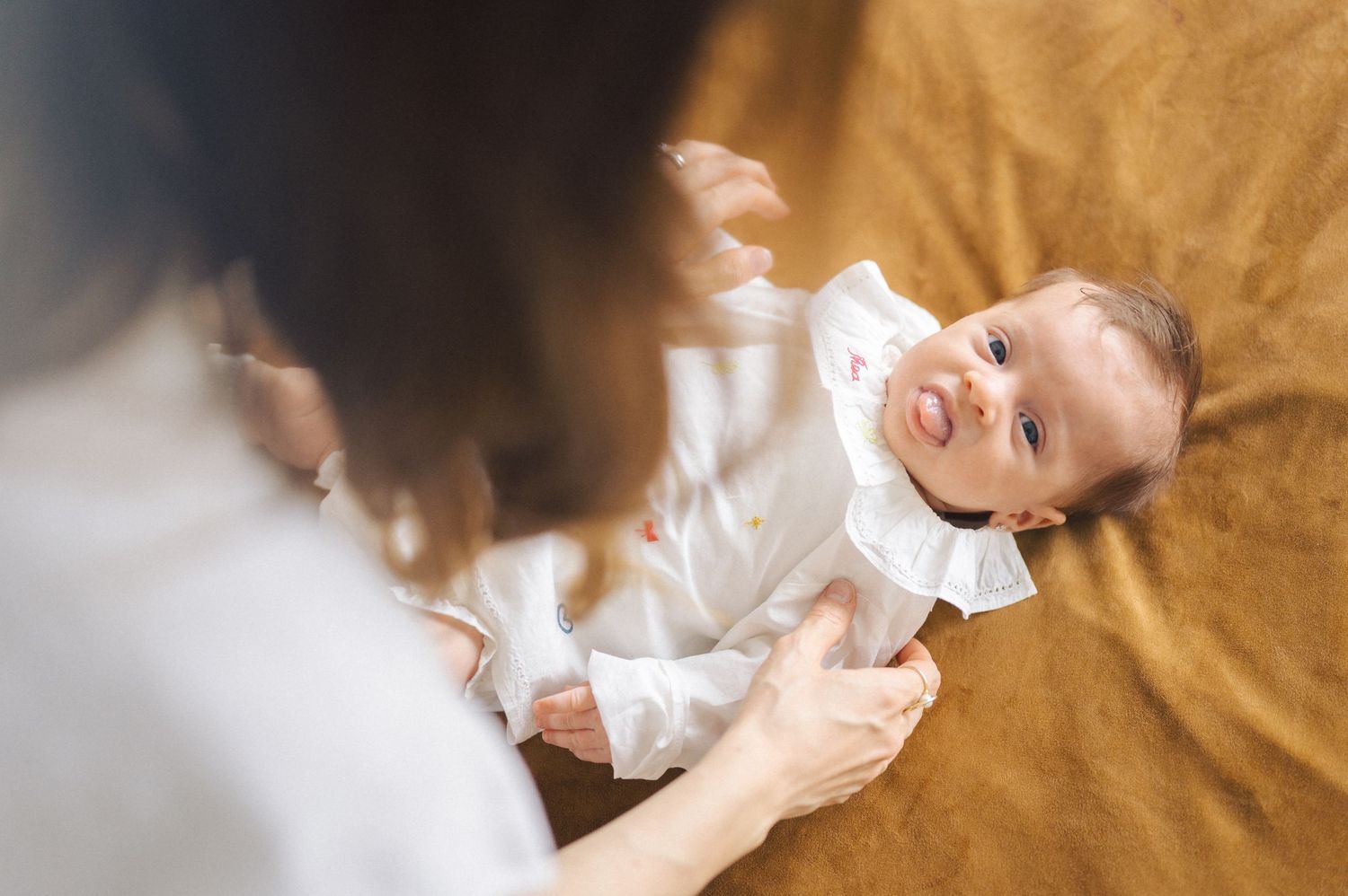 Baby wearing a white outfit lies on brown fabric while hands reach in from the side of the frame.
