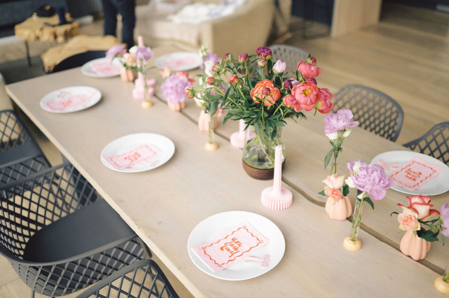 Table set with pink floral plates and coral flower centerpiece arrangements in glass vases on beige tablecloth.