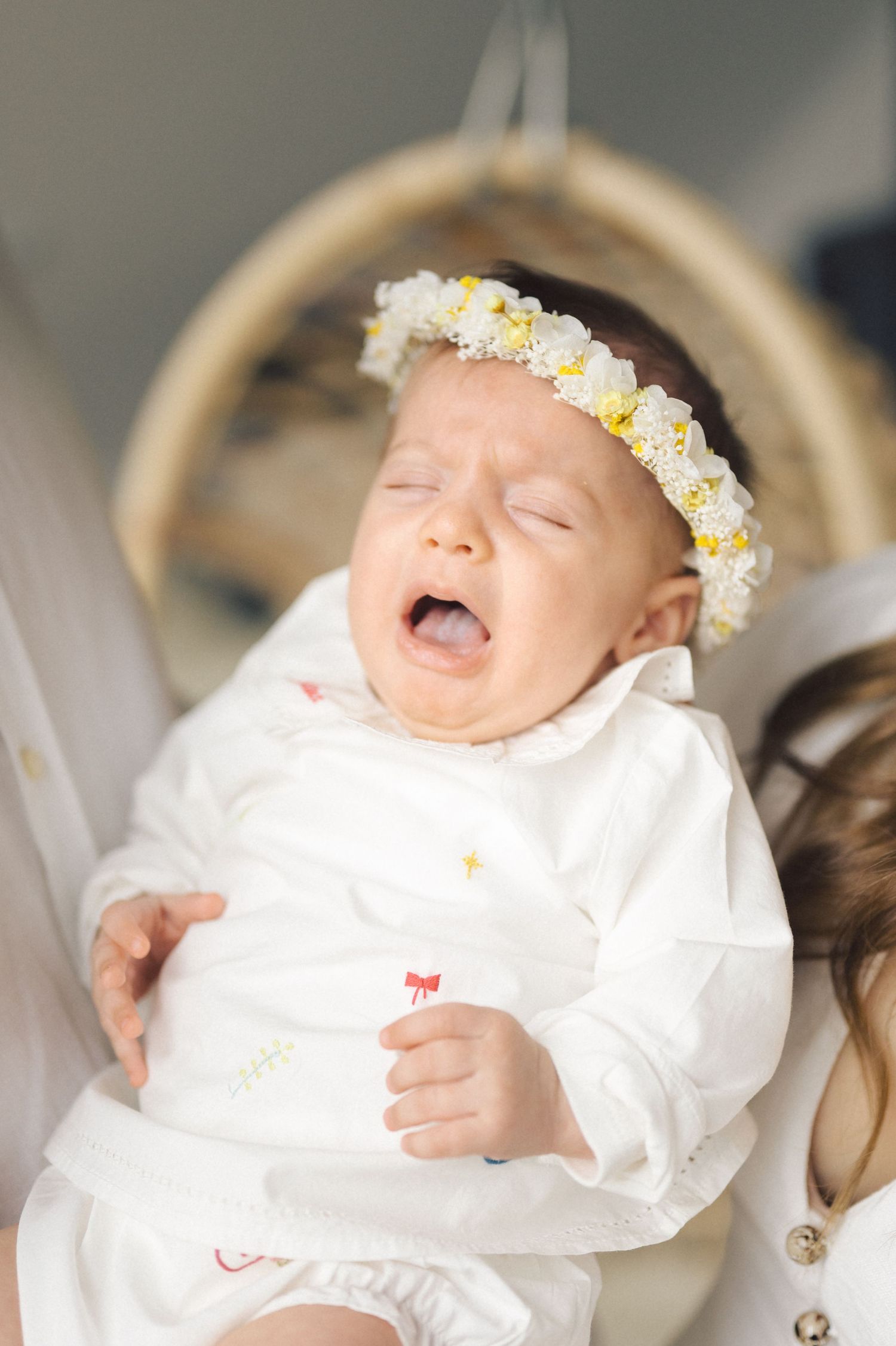 Baby wearing white dress and floral crown yawns while laying back during a special occasion.