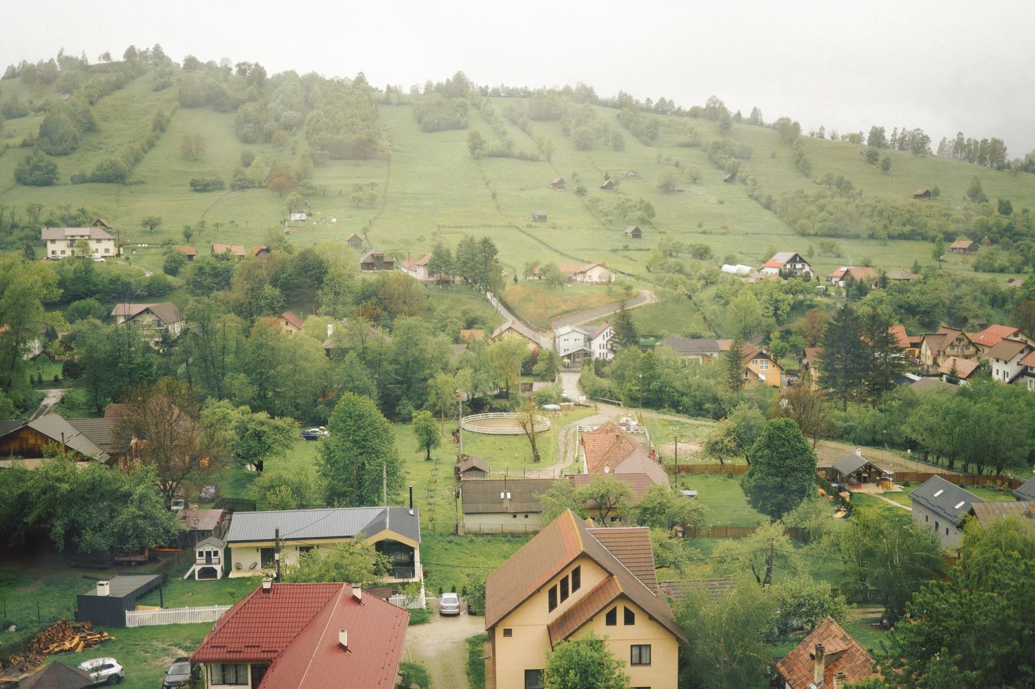 Aerial view of a rural hillside village with scattered houses and red roofs nestled among green slopes and trees.