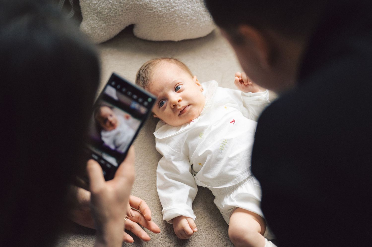 Baby lying on white surface while someone takes pictures with a smartphone camera.