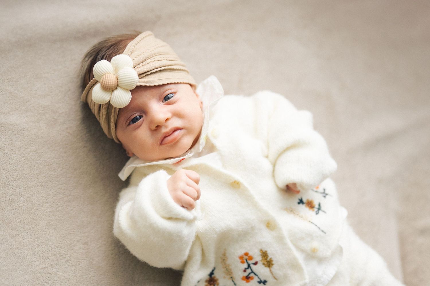 A peaceful baby wearing a white fleece outfit with a delicate floral headband rests on soft fabric.