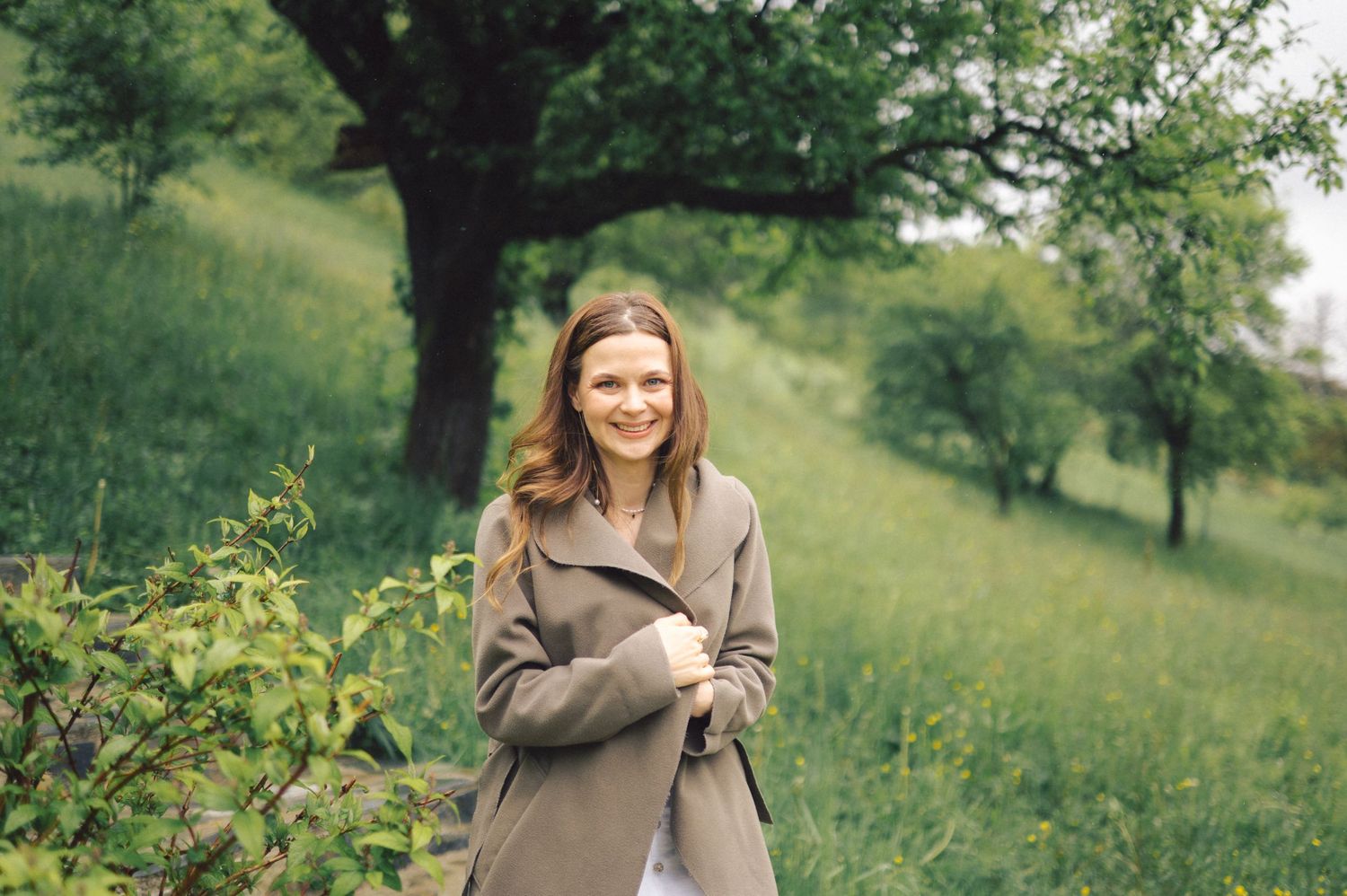 Person in beige coat smiling while walking in a lush green hillside on a sunny day.