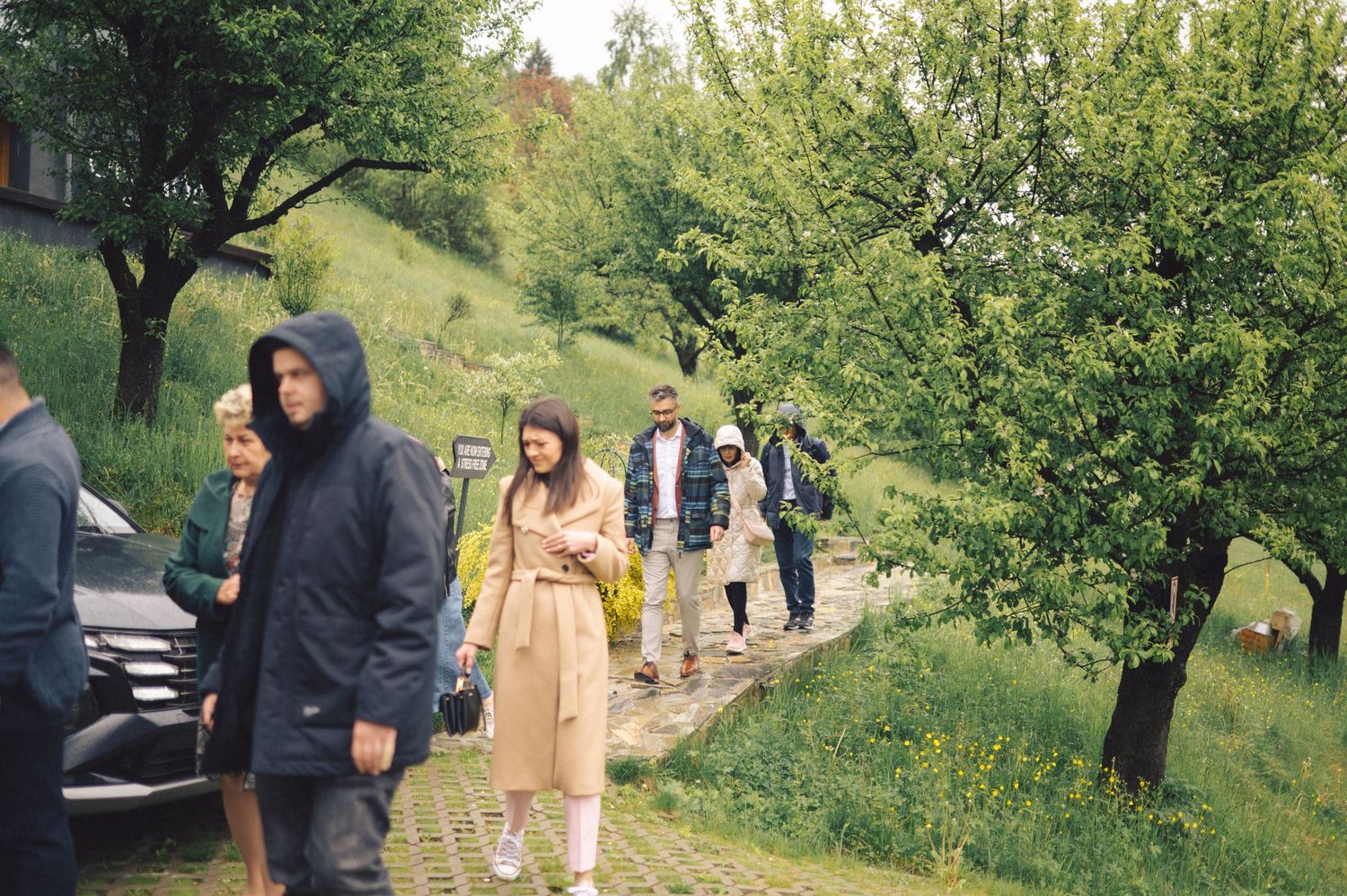 People in casual outerwear walking along a hillside path surrounded by green trees on an overcast day.
