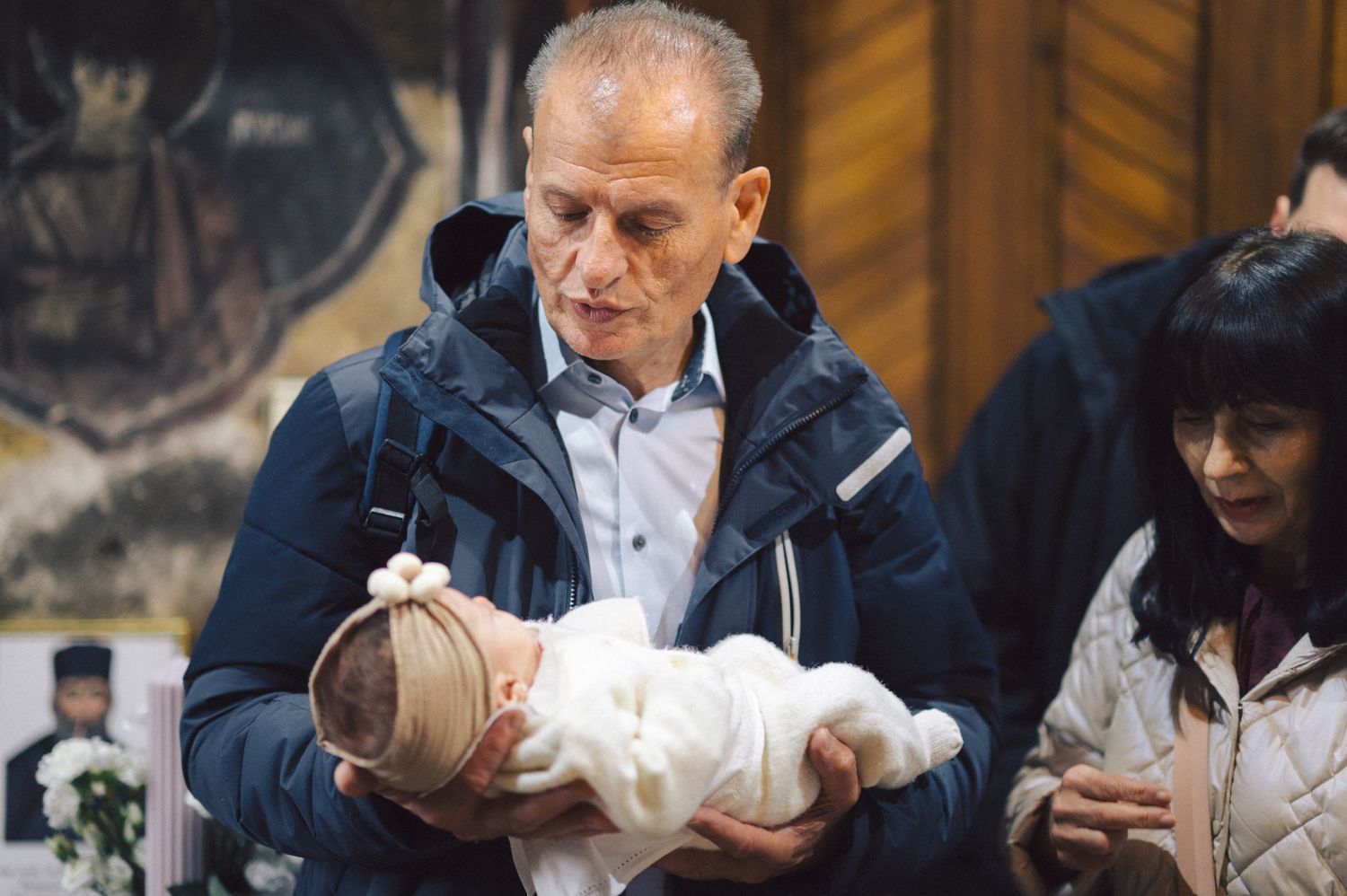 A person in a navy jacket tenderly holds a small baby wrapped in white blankets inside a building.