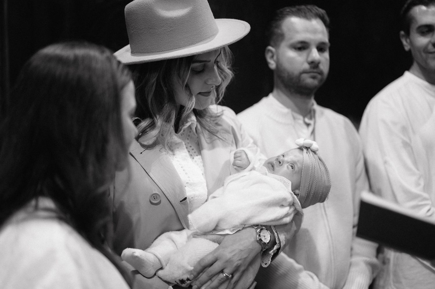 A black and white photo of a christening ceremony with adults in white clothing and someone wearing a gray hat.