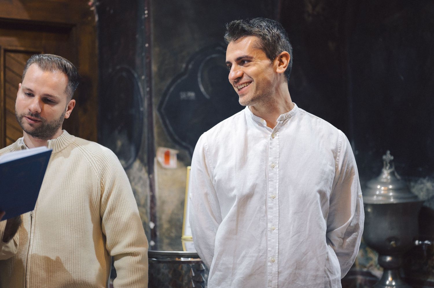 Two men stand together in a kitchen space, one wearing a beige sweater and one in a white button-up shirt, smiling.