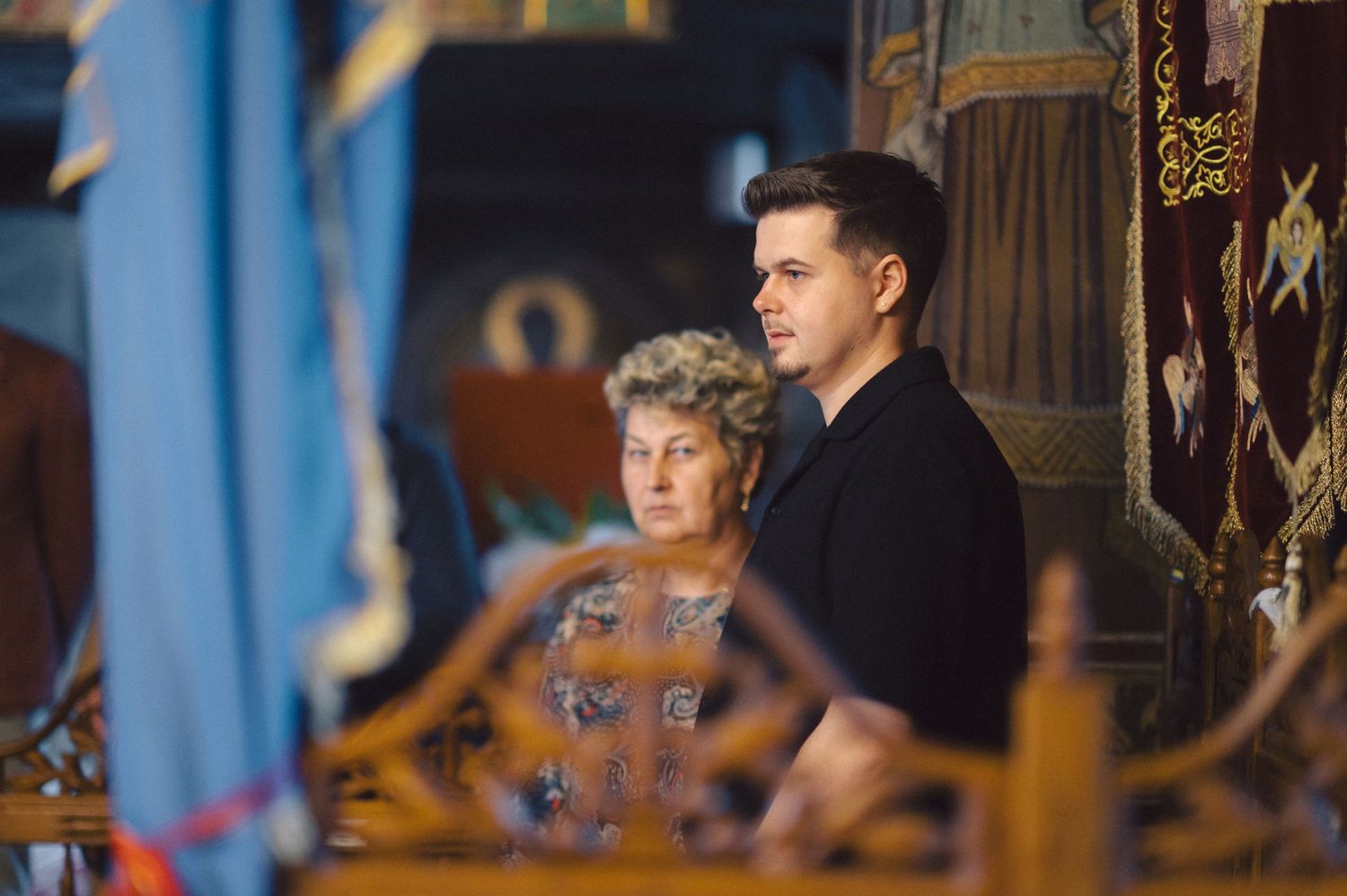 Two people in formal attire seated in an ornate, traditional church or cathedral setting with blue-tinted lighting.
