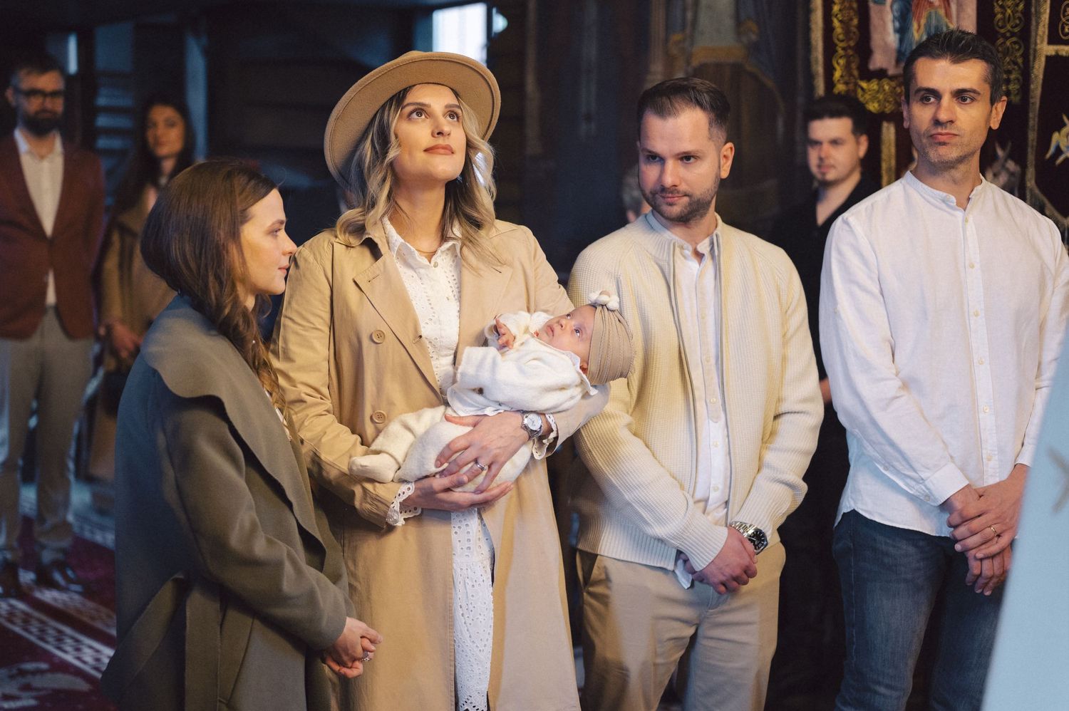 A group of people gather around a baby swaddled in pink being held by someone wearing a tan coat.
