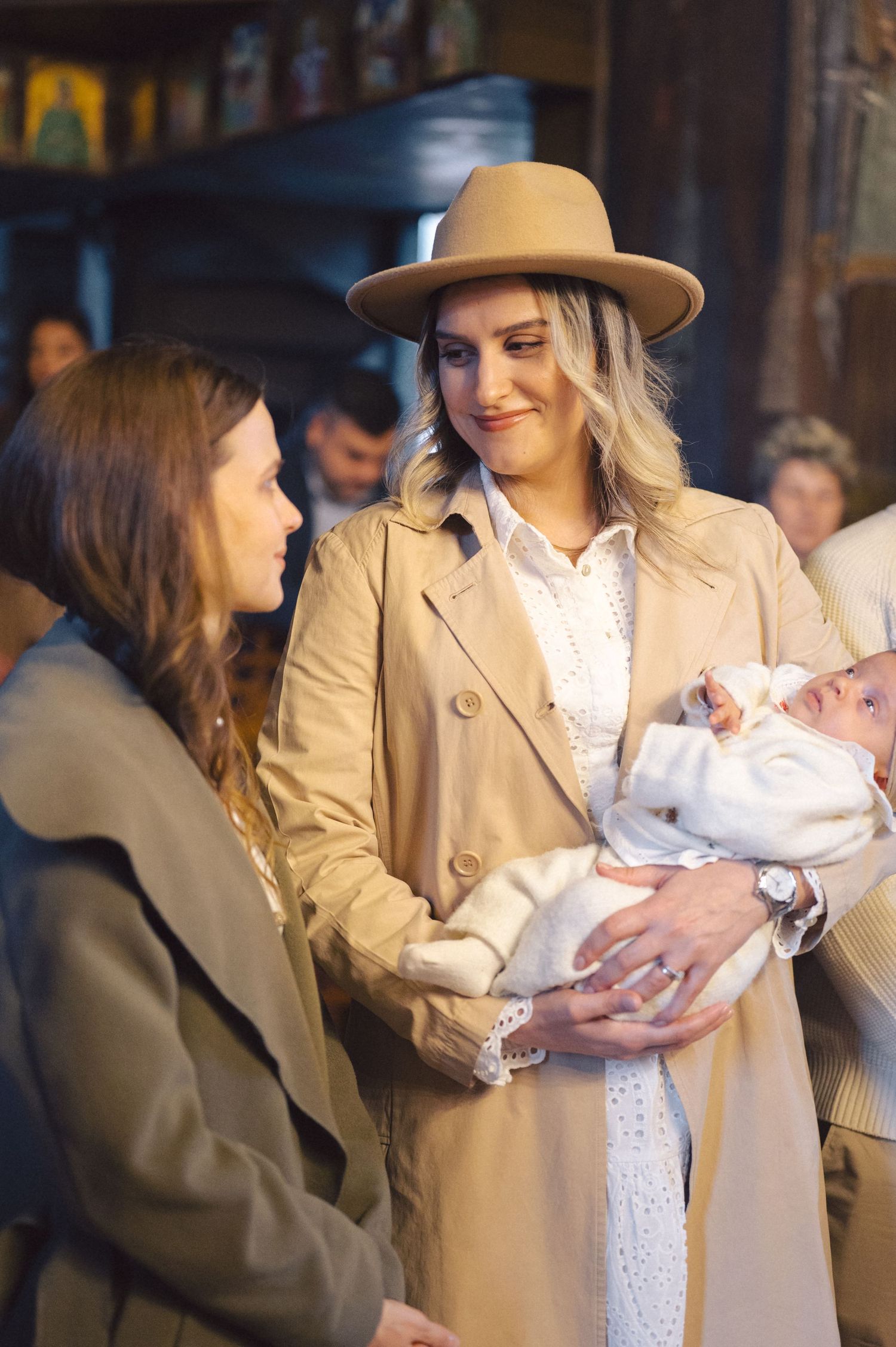 Two people having a warm conversation while holding a baby wrapped in white, with one person wearing a camel coat and hat.