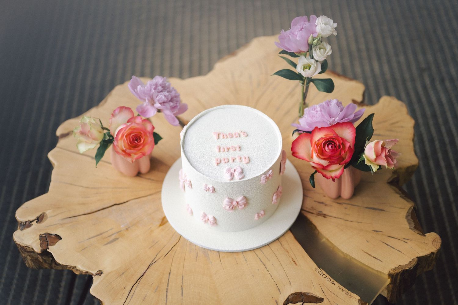 A white celebration cake surrounded by pink roses sits on a natural wood slice platter with floral decorations.