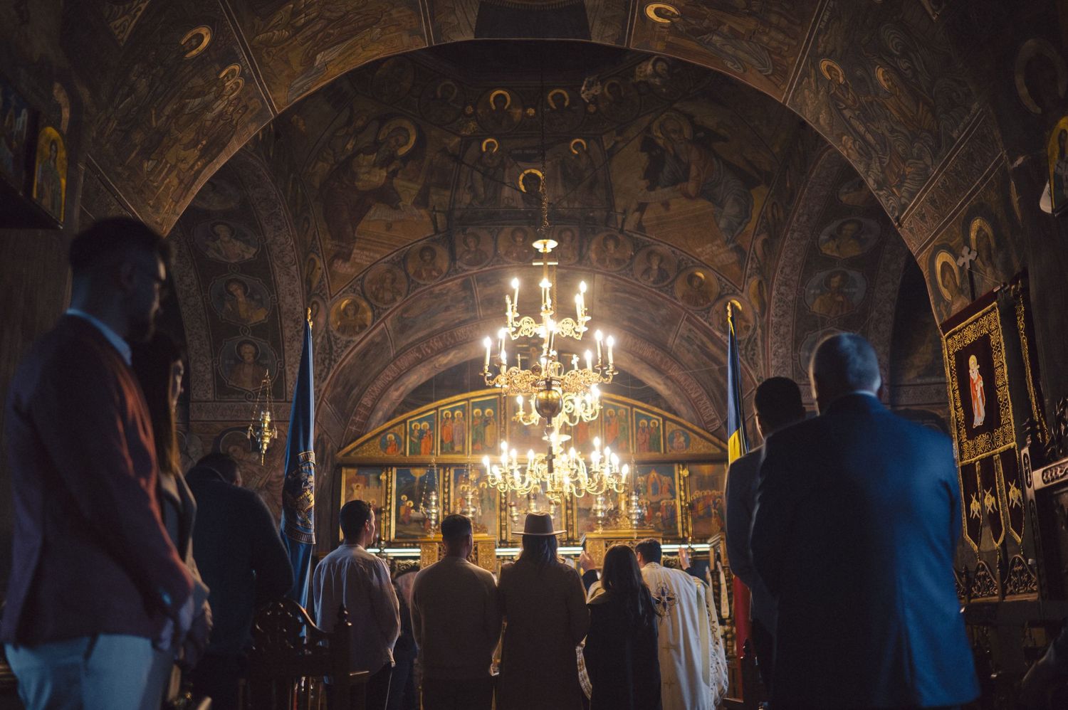 People stand inside an ornate Orthodox church with decorative frescoes and a glowing chandelier under vaulted ceilings.