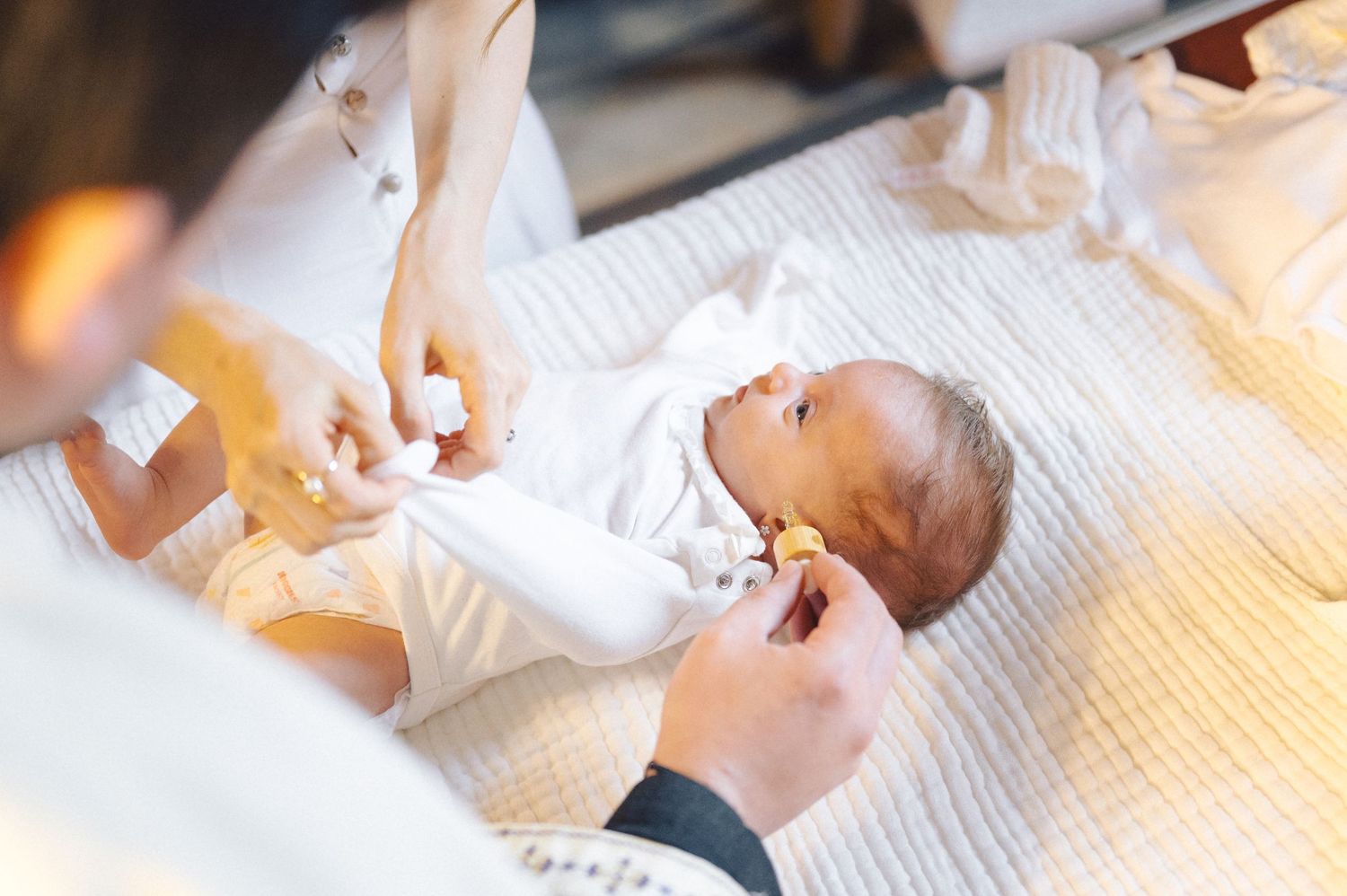 A small baby wearing white clothing lies on a white surface while being dressed by caregivers' hands.