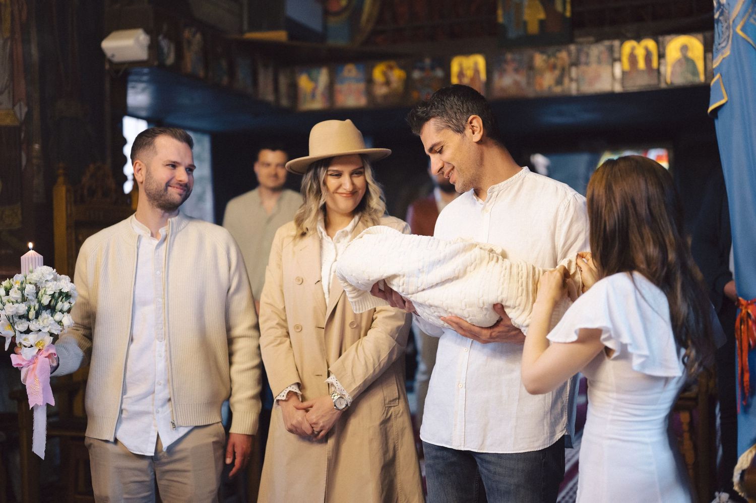 A group of people gather indoors to meet a newborn baby wrapped in white blankets.