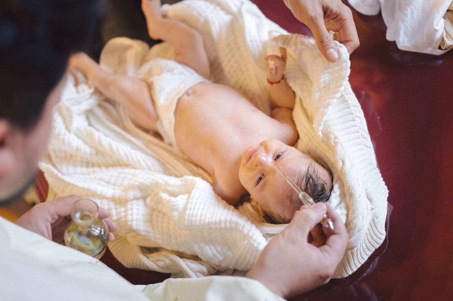 Baby lies on white blanket during diaper change with hands reaching to help from both sides.
