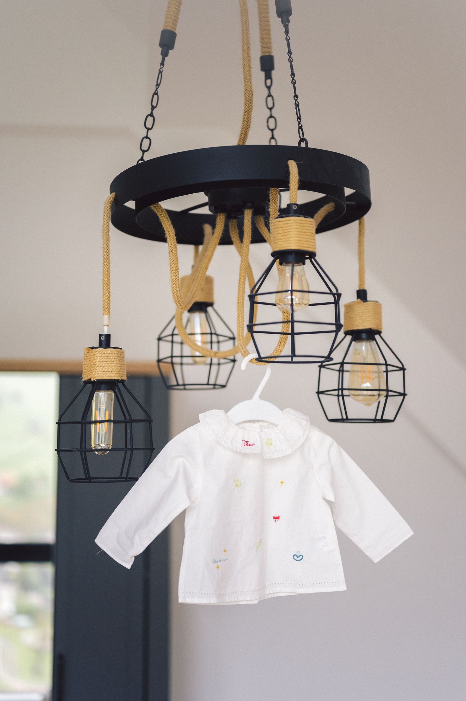 A modern ceiling light fixture with black metal cages and wooden accents hangs above a small white baby jacket.