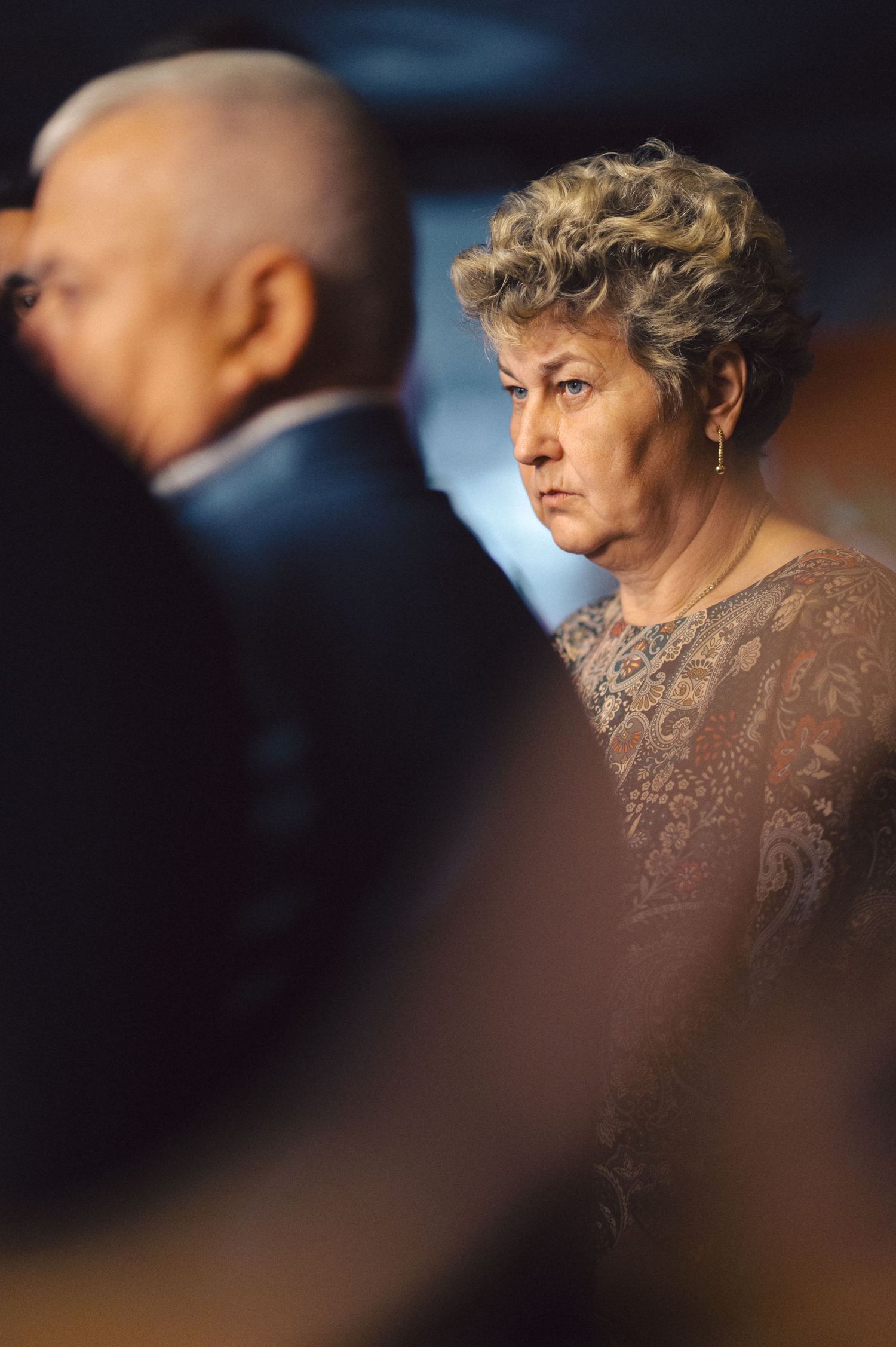 Person wearing a brown lace dress in soft lighting stands in contemplative profile against blurred background.