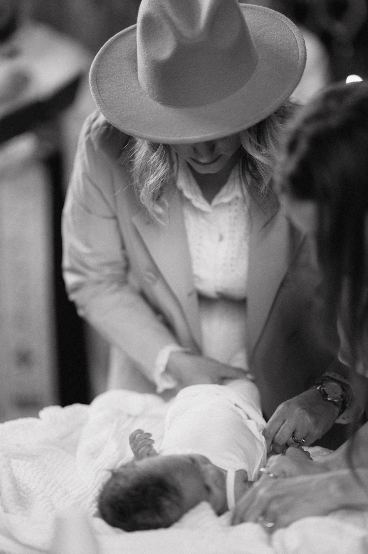 A person wearing a wide-brimmed hat and coat leans over to tend to a baby wrapped in white blankets in this black and white photo.