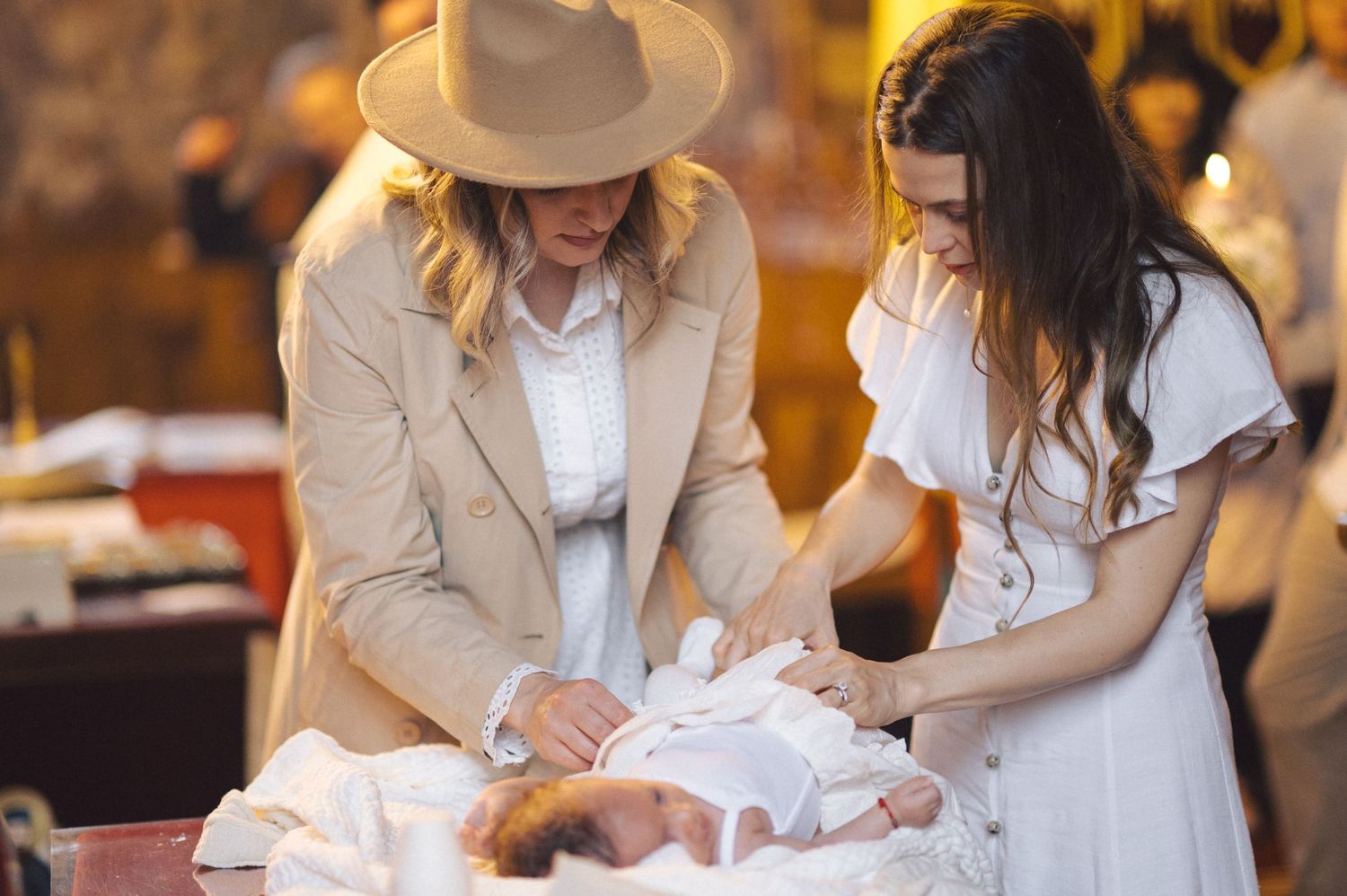 Two people in white clothing gently attend to a baby lying on a changing table in a warm-lit indoor setting.