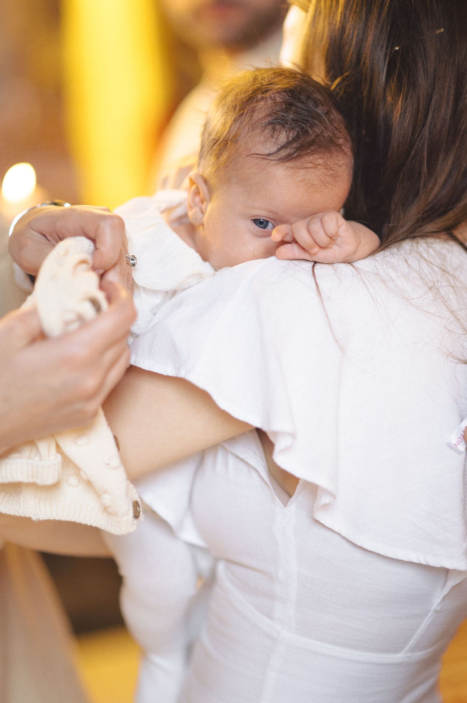A small baby in white clothing rests their head on a parent's shoulder while being held.