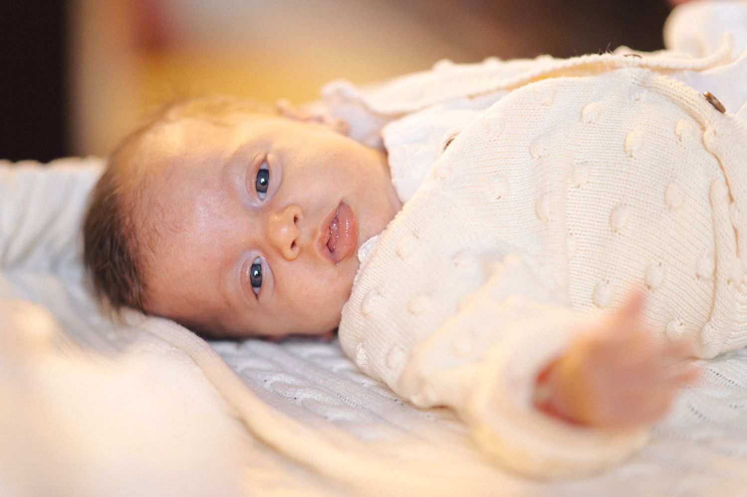 A baby lies on soft bedding wearing a light colored sleeper with bright eyes looking at the camera.