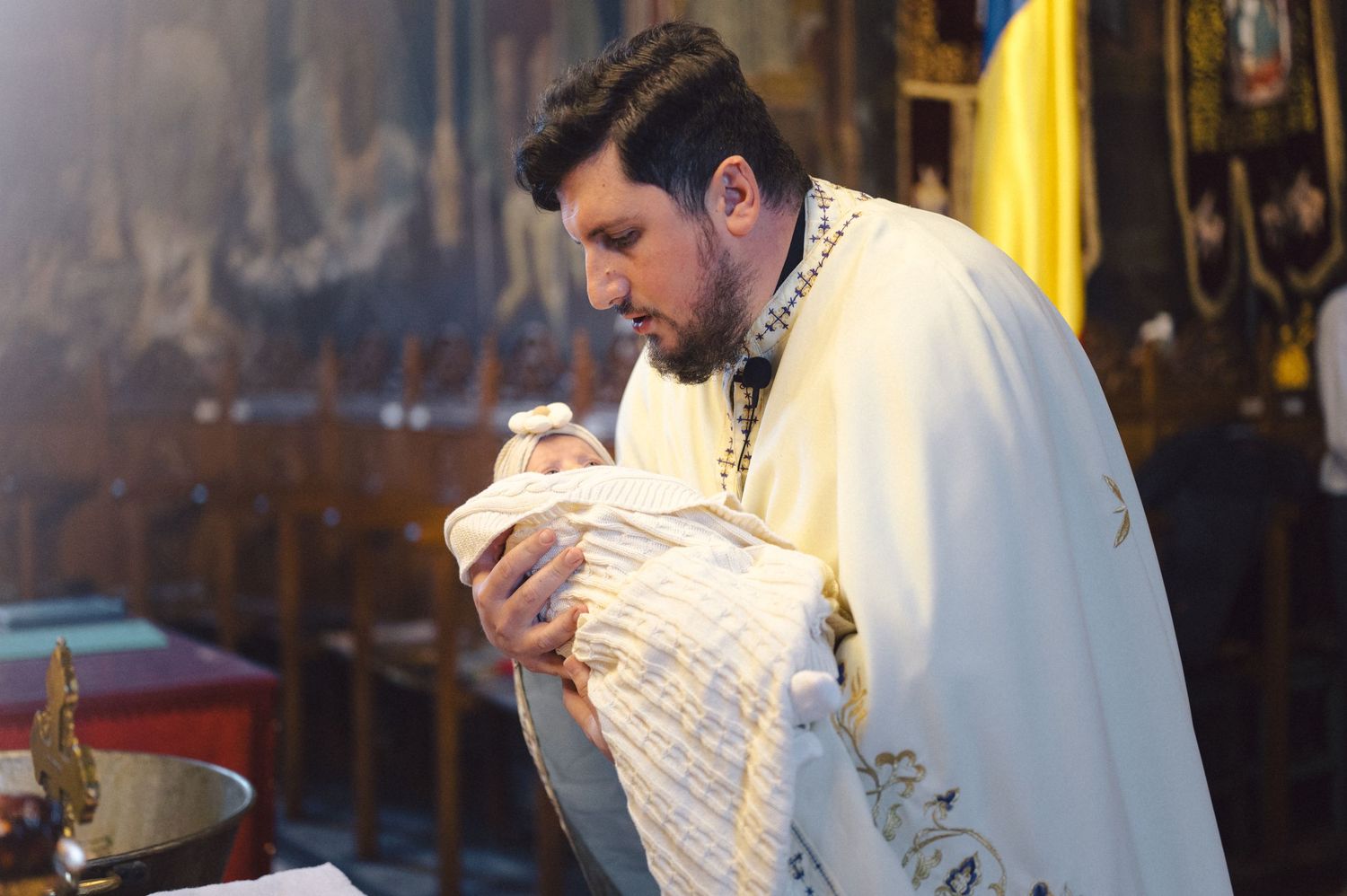 Orthodox priest wearing white vestments performs a baptism ceremony in a church amid incense smoke and candles.