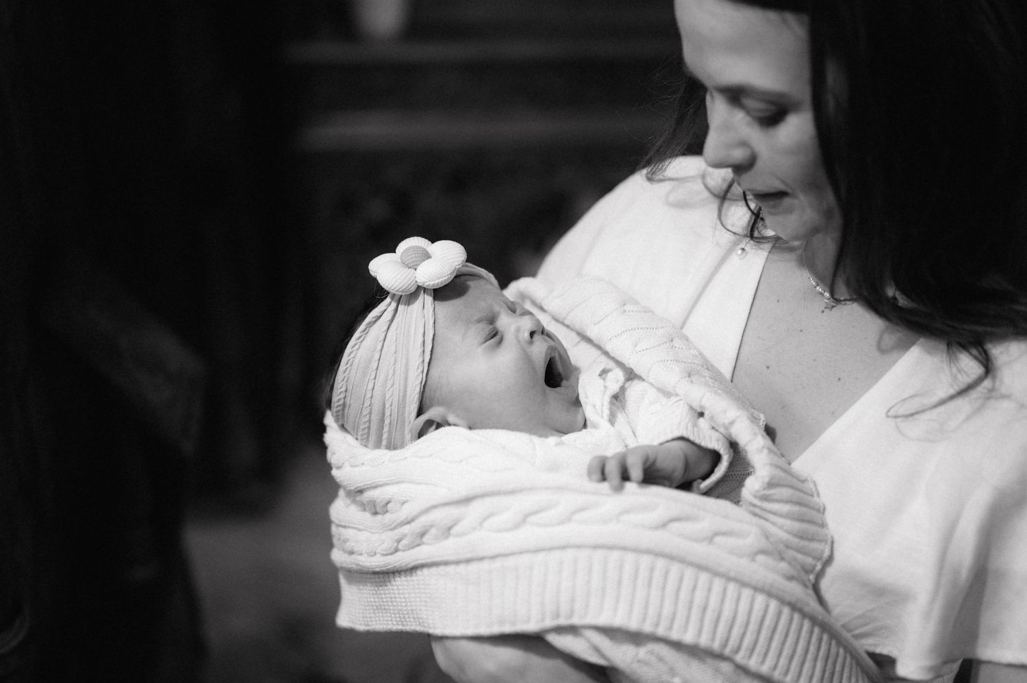 A tender black and white photograph shows a parent lovingly cradling their swaddled newborn baby wrapped in white blankets.
