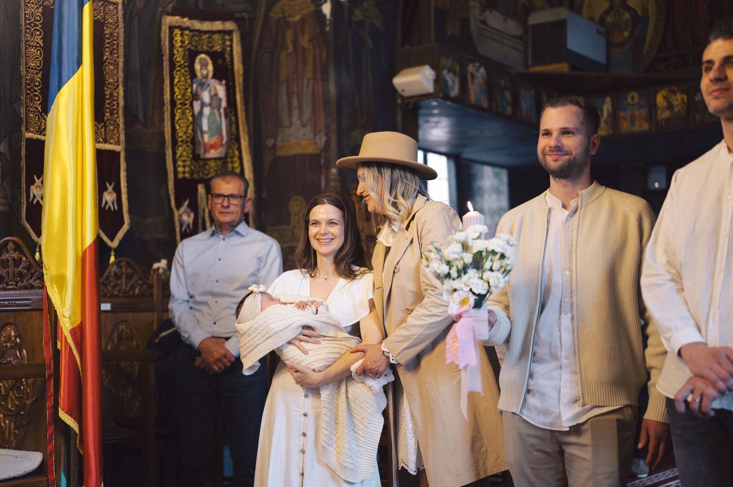 A group of people gather inside a church for what appears to be a christening ceremony with Romanian flag visible.
