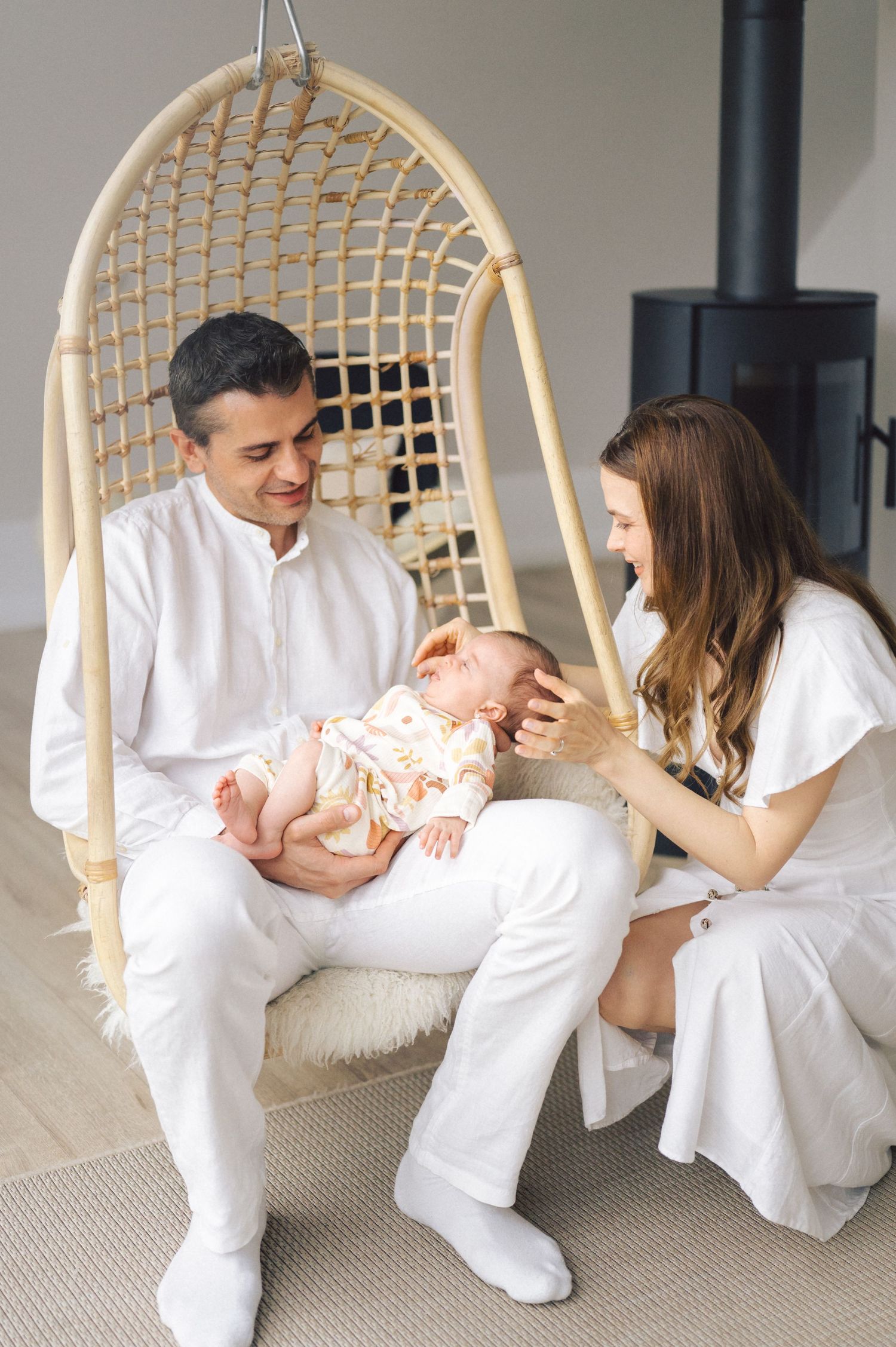 A couple dressed in white sits in a hanging rattan chair while holding a baby in a nursery setting with a wood stove.