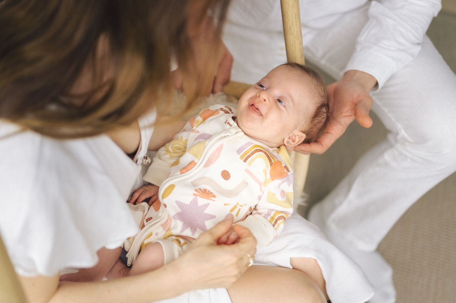 A baby wearing a peach-patterned onesie lies on a white surface while being gently held.