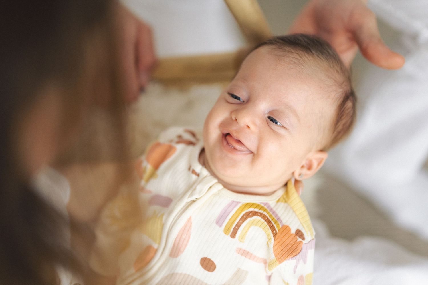 A baby wearing a rainbow print onesie smiles joyfully while lying on white bedding.
