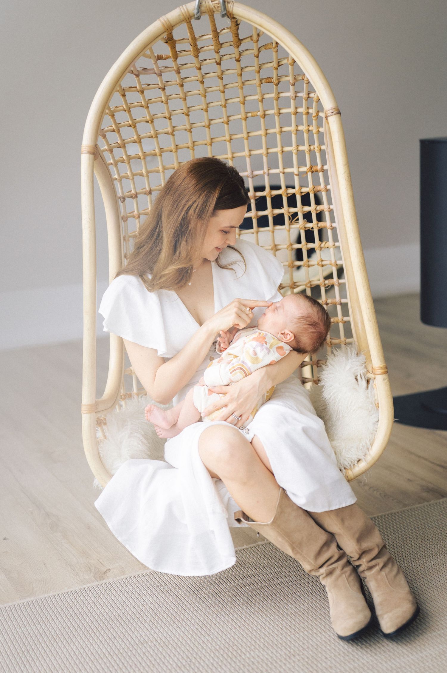A tender moment captured in a hanging rattan chair as a parent in a white dress cradles a sleeping baby.