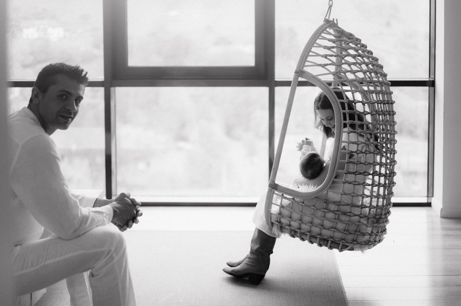 A wicker hanging chair by a window holds a person while someone sits nearby in a minimalist interior.