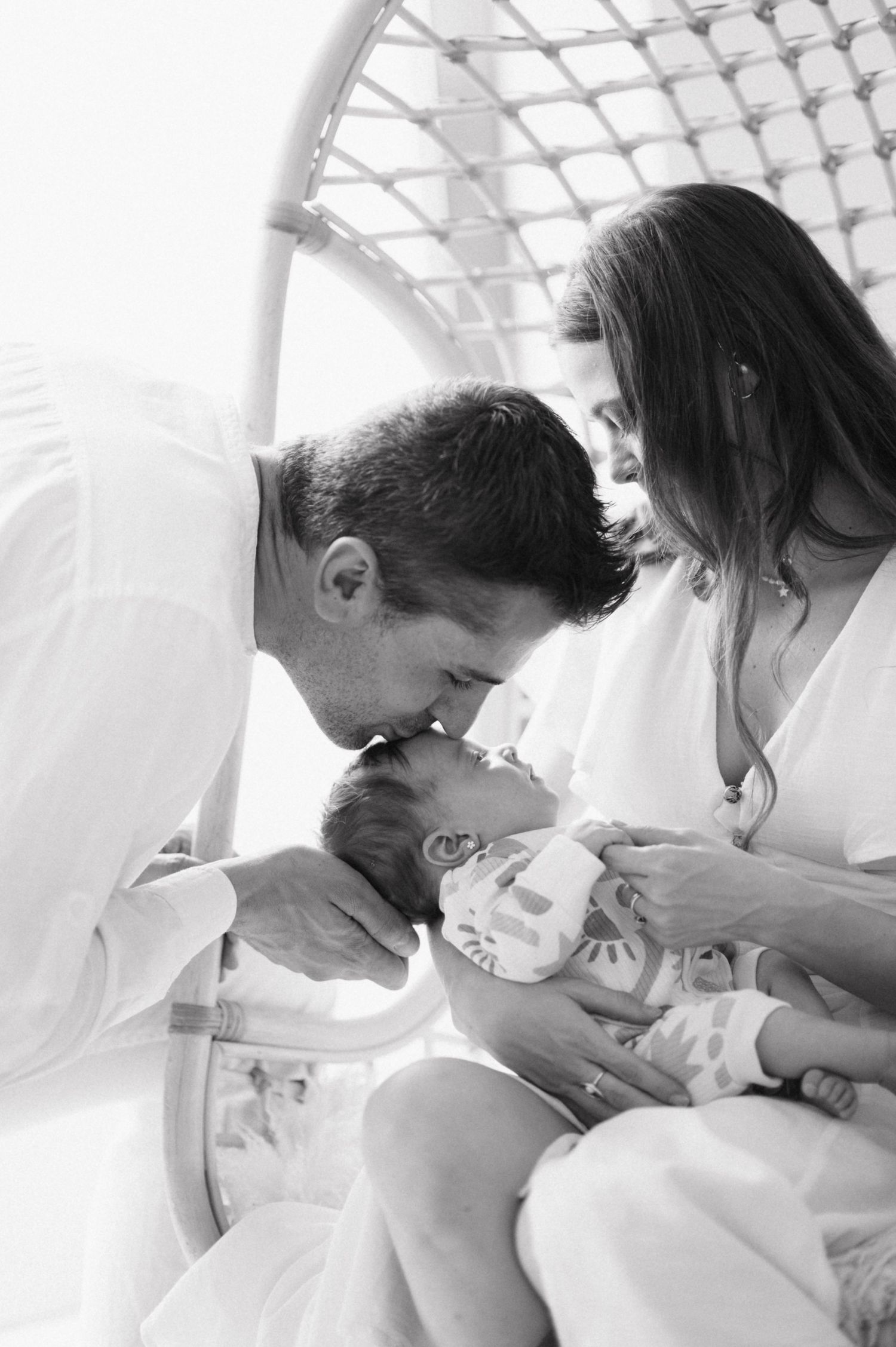 A tender black and white family moment captured under a geometric ceiling structure as parents lovingly gaze at their baby.