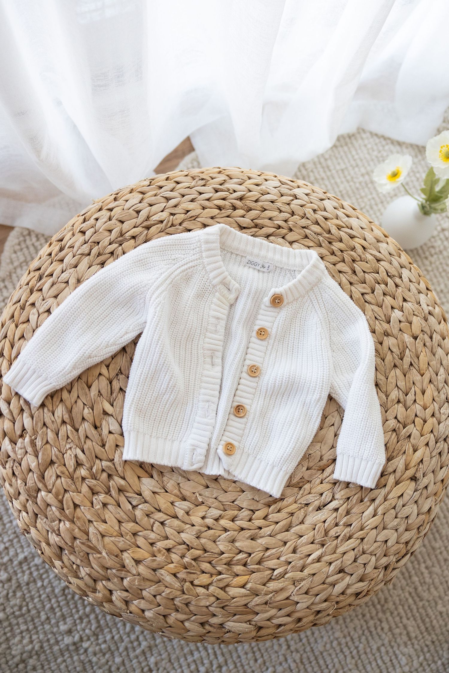 White knitted baby cardigan sweater with wooden buttons displayed on round woven straw mat with floral decor.